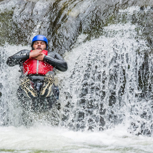 A man wearing a wetsuit, hard hat and buoyancy aid slides down a waterfall