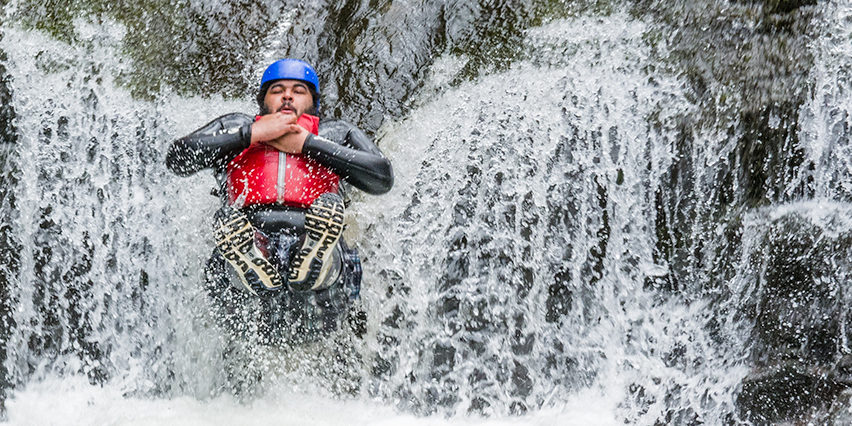 CalloftheWild-1 A man wearing a wetsuit, hard hat and buoyancy aid slides down a waterfall