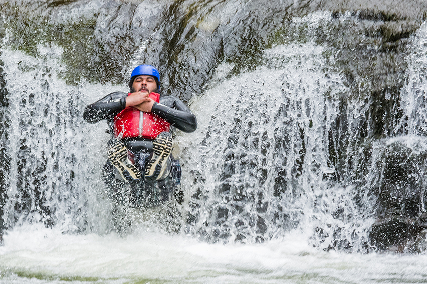 A man wearing a wetsuit, hard hat and buoyancy aid slides down a waterfall